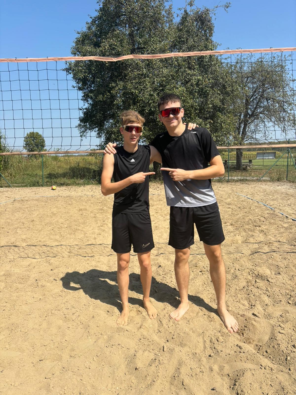 The image depicts two men standing on a sandy volleyball court, posing for a photograph. They are both wearing black athle...